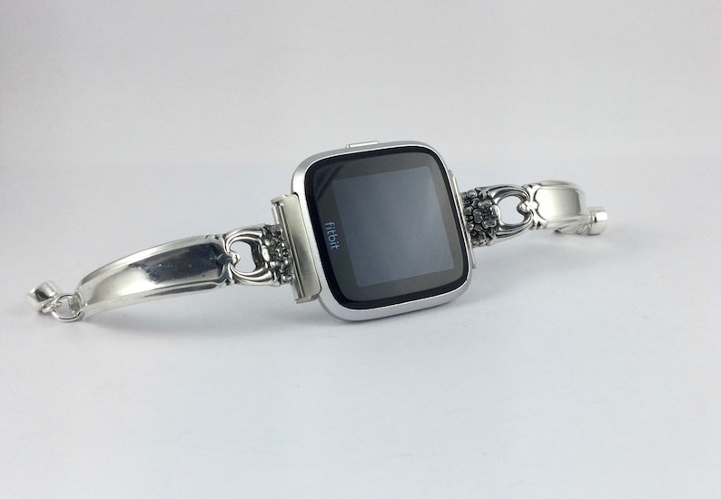 May include: A silver Fitbit smartwatch with a black screen is attached to a silver bracelet made from a repurposed silverware handle.