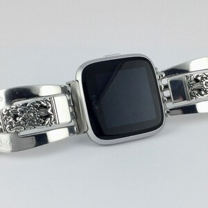 May include: Silver watch band with an ornate design. The band is made of two pieces that are connected by a buckle. The watch face is black and has the word "Fitbit" on it.