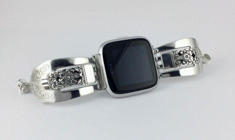 May include: Silver watch band with an ornate design. The band is made of two pieces that are connected by a buckle. The watch face is black and has the word "Fitbit" on it.