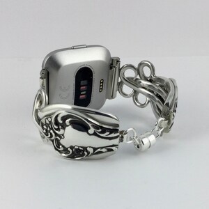May include: A silver smartwatch with a silver spoon bracelet. The watch face is square and has a black screen with white text that reads "CE".