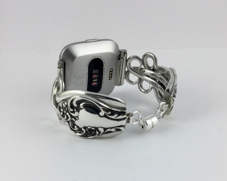 May include: A silver smartwatch with a silver spoon bracelet. The watch face is square and has a black screen with white text that reads "CE".