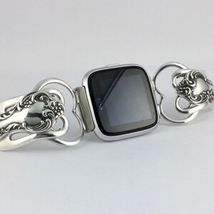 Puede incluir: Un reloj inteligente Fitbit plateado con una pantalla negra est&aacute; conectado a una pulsera plateada hecha de dos cucharas. Las cucharas est&aacute;n dobladas en forma de coraz&oacute;n y tienen dise&ntilde;os ornamentados.