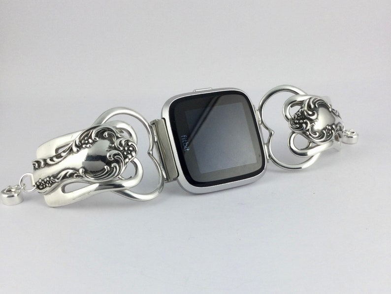 Puede incluir: Un reloj inteligente Fitbit plateado con una pantalla negra est&aacute; conectado a una pulsera plateada hecha de dos cucharas. Las cucharas est&aacute;n dobladas en forma de coraz&oacute;n y tienen dise&ntilde;os ornamentados.