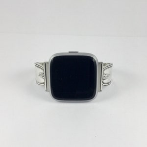 May include: A silver watch band made from a repurposed silverware piece. The band is designed to fit a square-shaped smartwatch.