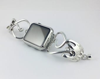 Puede incluir: Un reloj inteligente de tono plateado con pantalla rectangular y una correa de metal decorativa. La correa presenta intrincados patrones de metal calado y en espiral en ambos lados, con un sutil grabado floral y un pequeño cierre magnético.