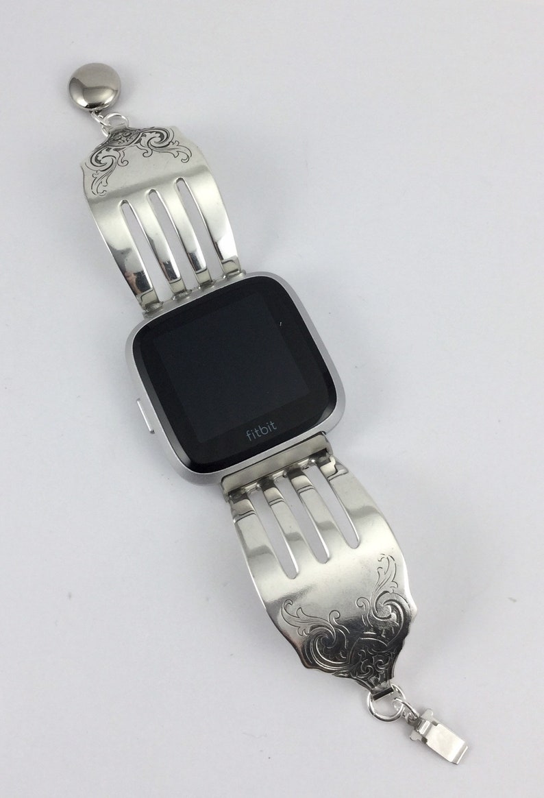 Pu&ograve; includere: Un orologio intelligente Fitbit argentato con uno schermo nero &egrave; attaccato a un braccialetto a forchetta argentato. Il braccialetto &egrave; composto da due forchette con i denti rivolti l'uno verso l'altro. Il braccialetto presenta un design decorativo sulla parte superiore e inferiore delle forchette.