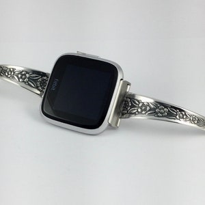 Puede incluir: Un reloj inteligente Fitbit plateado con una banda con estampado floral. La banda est&aacute; hecha de una pieza de cubiertos reutilizada.