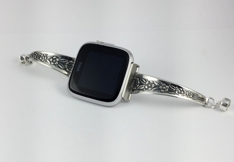 Puede incluir: Un reloj inteligente Fitbit plateado con una banda con estampado floral. La banda est&aacute; hecha de una pieza de cubiertos reutilizada.