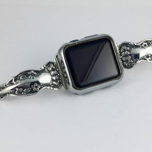 May include: A silver-toned smartwatch featuring a rectangular black screen and a metallic watch band. The band is crafted from repurposed silverware, displaying an intricate embossed floral pattern with small blossoms and leaves along the surface.