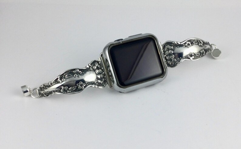 May include: A silver-toned smartwatch featuring a rectangular black screen and a metallic watch band. The band is crafted from repurposed silverware, displaying an intricate embossed floral pattern with small blossoms and leaves along the surface.