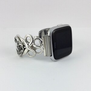 May include: Silver watch band with a decorative, ornate design. The band is attached to a silver Fitbit smartwatch with a black screen.
