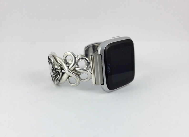 May include: Silver watch band with a decorative, ornate design. The band is attached to a silver Fitbit smartwatch with a black screen.