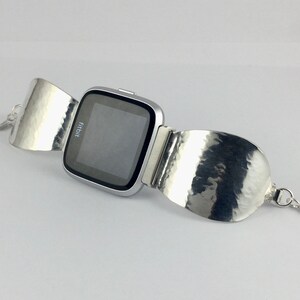 May include: A silver Fitbit smartwatch with a hammered silver band. The band is designed to wrap around the wrist and has a small heart-shaped charm.
