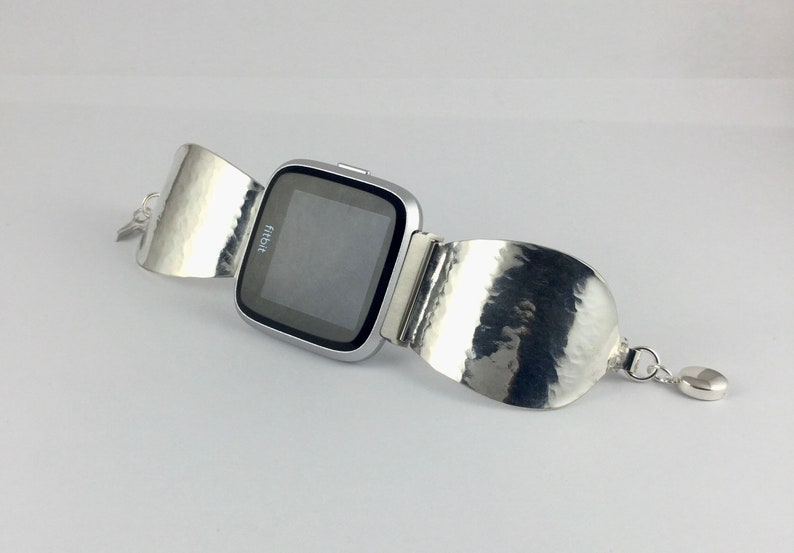 May include: A silver Fitbit smartwatch with a hammered silver band. The band is designed to wrap around the wrist and has a small heart-shaped charm.