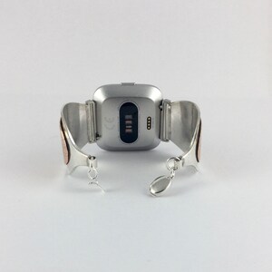 May include: Silver and copper bracelet with a silver smartwatch attached. The smartwatch has a black screen with the CE mark and a row of four small lights.