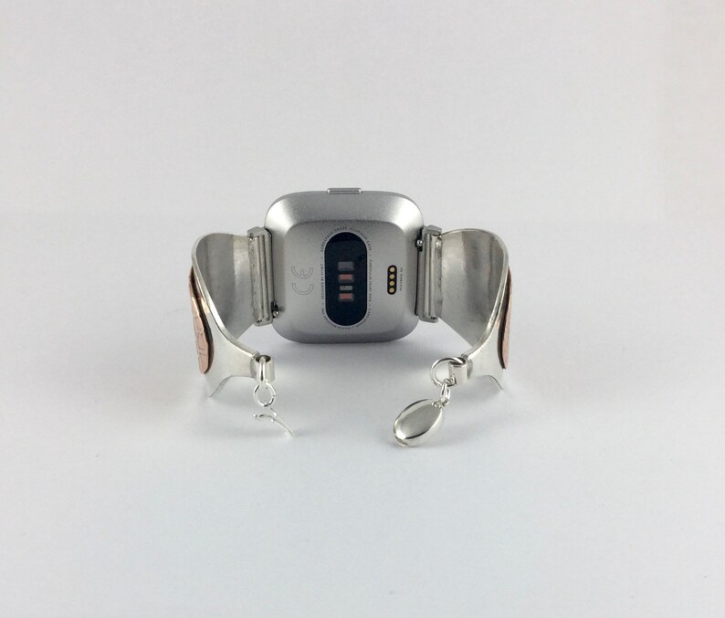 May include: Silver and copper bracelet with a silver smartwatch attached. The smartwatch has a black screen with the CE mark and a row of four small lights.