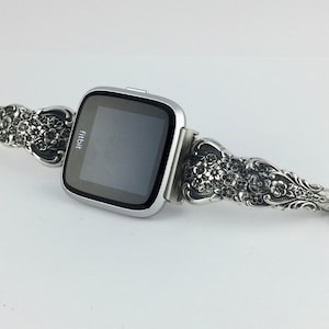 May include: Silver Fitbit smartwatch with a decorative band. The band features an ornate floral design. The watch face is black with the Fitbit logo. The band is made of a silver-colored metal.