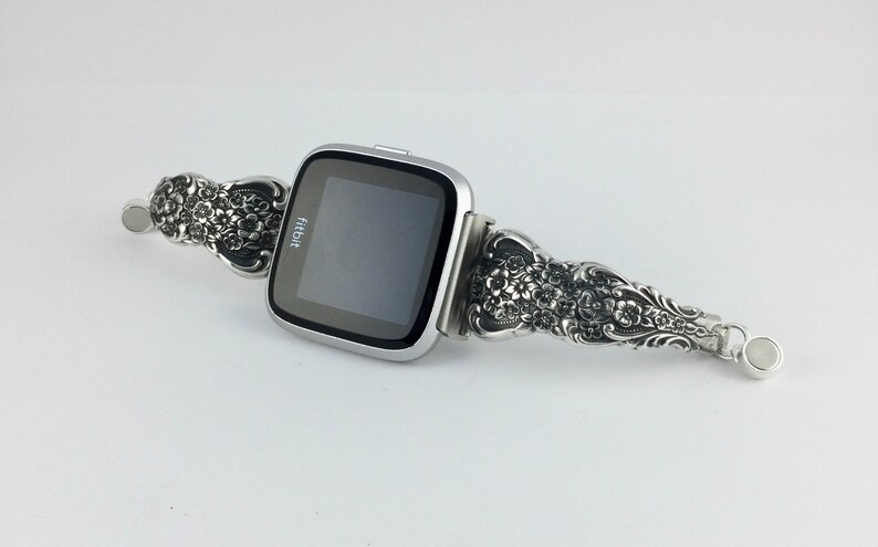 May include: Silver Fitbit smartwatch with a decorative band. The band features an ornate floral design. The watch face is black with the Fitbit logo. The band is made of a silver-colored metal.