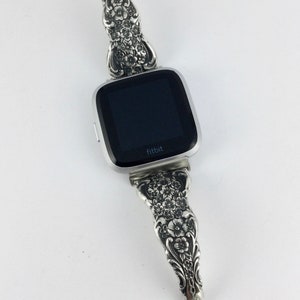 May include: Silver Fitbit watch with a decorative band. The band is made from ornate silverware with a floral design. The watch face is black with the brand name "fitbit" in white. The band is connected with a small silver clasp.