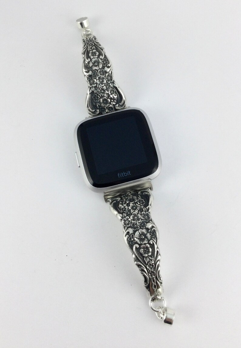 May include: Silver Fitbit watch with a decorative band. The band is made from ornate silverware with a floral design. The watch face is black with the brand name "fitbit" in white. The band is connected with a small silver clasp.