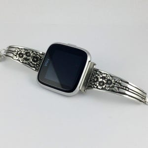 Pu&ograve; includere: Un orologio intelligente Fitbit argentato con un cinturino a motivo floreale realizzato con posate riciclate.