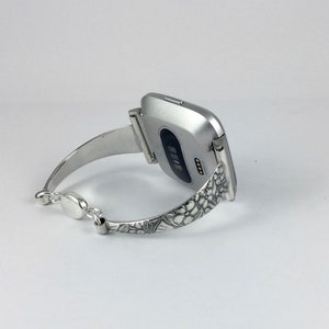 May include: Silver smartwatch with a floral patterned band. The watch face is silver and has a black screen.