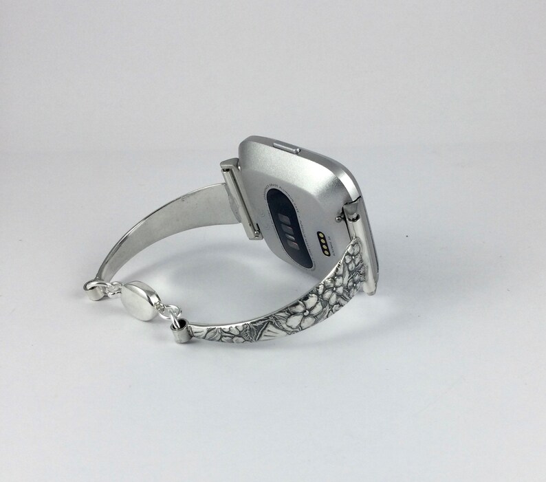 May include: Silver smartwatch with a floral patterned band. The watch face is silver and has a black screen.