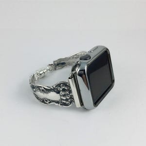 May include: A silver-toned smartwatch with a polished metallic finish and a black screen. The watch band is crafted from repurposed silver-colored metal cutlery featuring an intricate embossed floral pattern along the entire length of the strap.