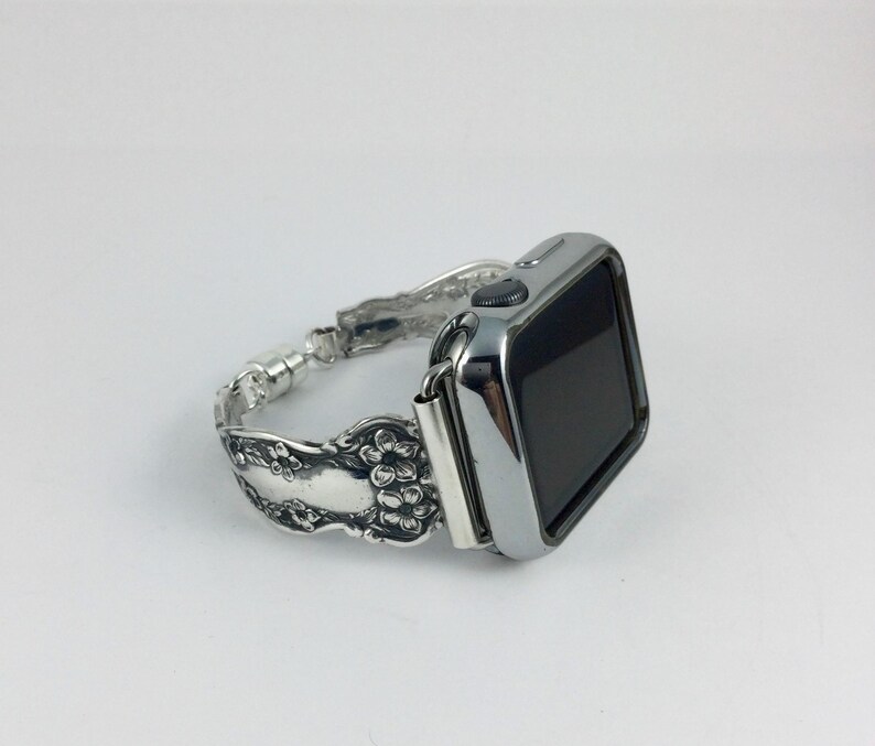 May include: A silver-toned smartwatch with a polished metallic finish and a black screen. The watch band is crafted from repurposed silver-colored metal cutlery featuring an intricate embossed floral pattern along the entire length of the strap.