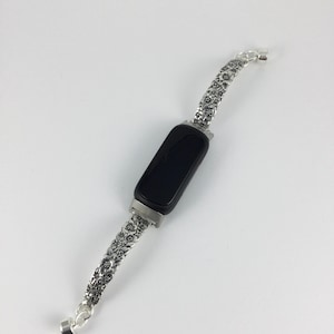 May include: A black fitness tracker with a silver floral patterned band. The band is made of a series of small, detailed flower designs. The tracker has a rectangular black screen and a silver clasp.