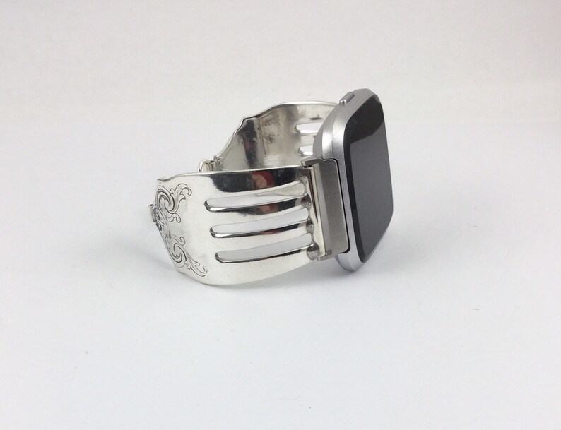 Pu&ograve; includere: Un cinturino per orologio in argento realizzato da una forchetta riutilizzata. Il cinturino presenta un design decorativo ed &egrave; attaccato a uno smartwatch argentato.