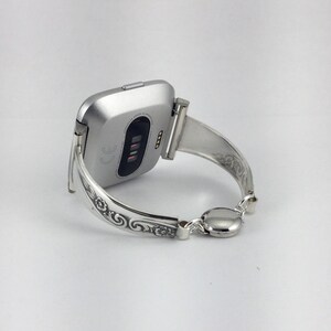 May include: Silver watch band with an ornate design. The band is a bangle style and has a clasp closure.