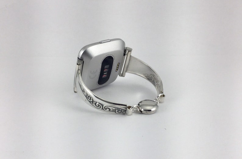May include: Silver watch band with an ornate design. The band is a bangle style and has a clasp closure.