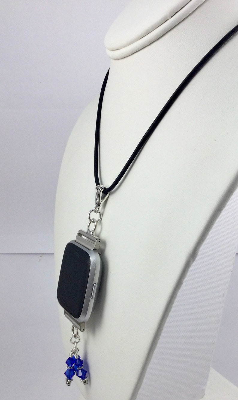 May include: A silver smartwatch pendant necklace with a black screen, suspended from a black cord. The pendant features a cluster of blue crystal beads. The necklace is displayed on a white mannequin.