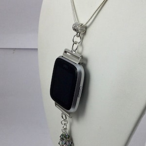 K&ouml;nnte beinhalten: Eine silberne Halskette mit einem silbernen Uhrenzifferblatt-Anh&auml;nger und einer silbernen Kette. Das Zifferblatt der Uhr ist schwarz und die Halskette hat einen silbernen Anh&auml;nger mit schillernden Kristallen.