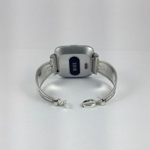 May include: Silver bracelet with a silver smartwatch attached. The bracelet has an ornate design and a clasp closure.