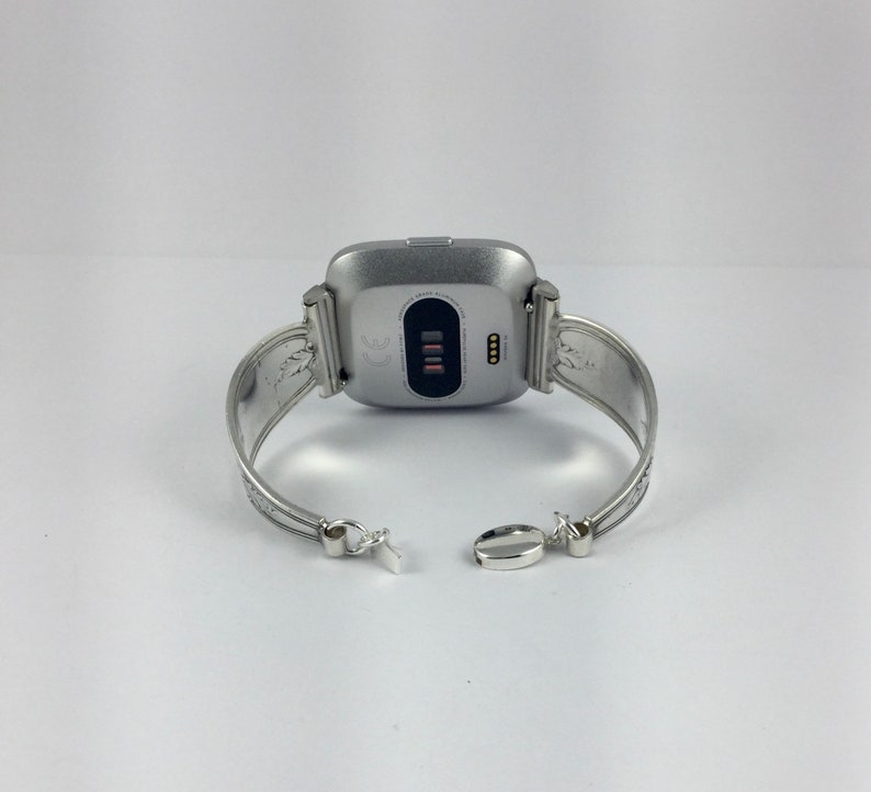 May include: Silver bracelet with a silver smartwatch attached. The bracelet has an ornate design and a clasp closure.