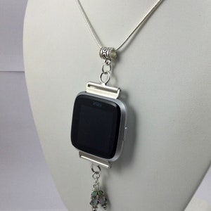 K&ouml;nnte beinhalten: Eine silberne Kettenkette mit einem silbernen Anh&auml;nger, der eine silberne Fitbit-Uhr h&auml;lt. Das Zifferblatt der Uhr ist schwarz und der Anh&auml;nger hat einen kleinen Kristall-Anh&auml;nger.