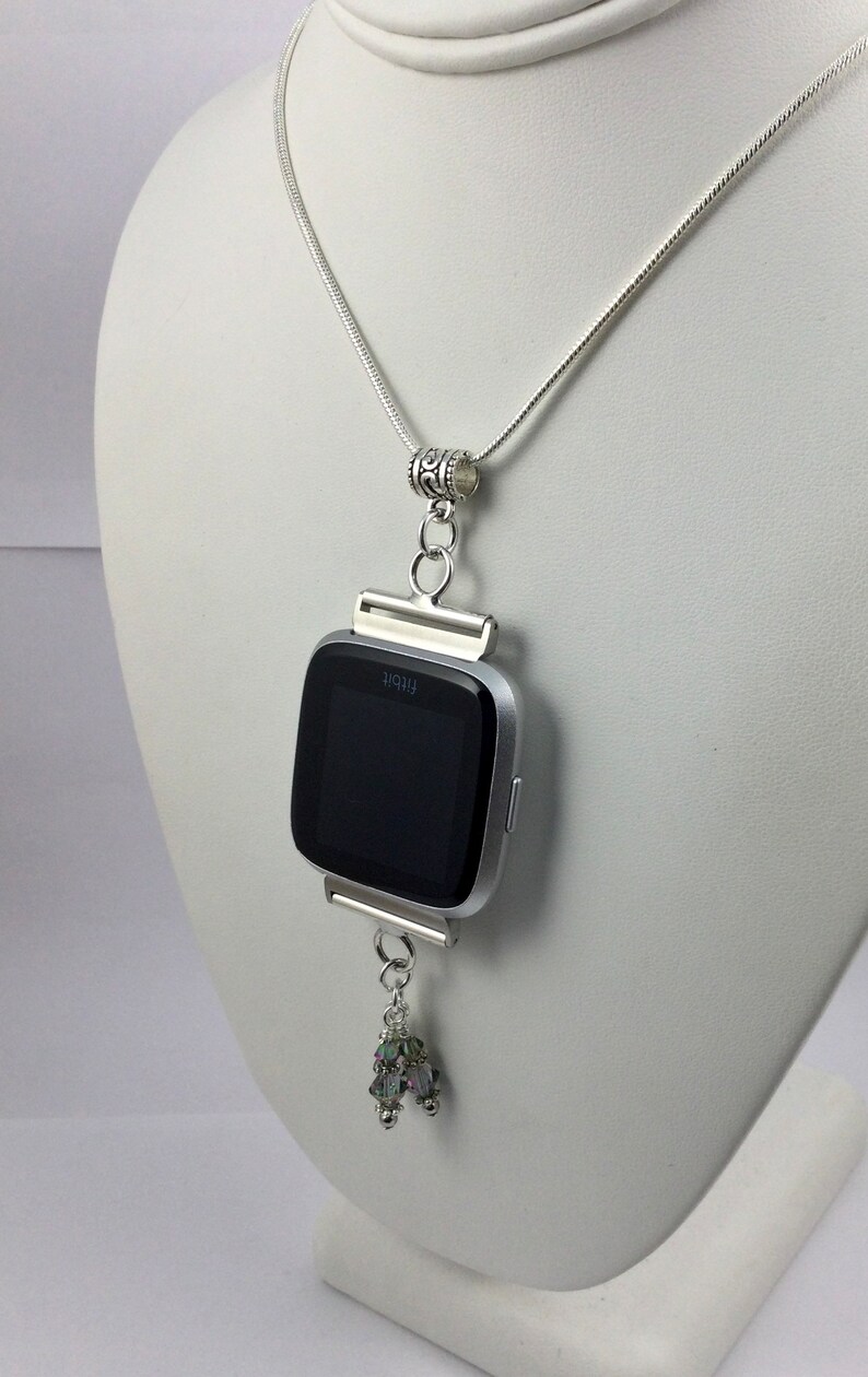 K&ouml;nnte beinhalten: Eine silberne Kettenkette mit einem silbernen Anh&auml;nger, der eine silberne Fitbit-Uhr h&auml;lt. Das Zifferblatt der Uhr ist schwarz und der Anh&auml;nger hat einen kleinen Kristall-Anh&auml;nger.