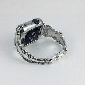 May include: A silver-toned smartwatch with a metallic, floral-patterned band. The watch features a rectangular case with a black circular sensor on the back and a side button. The band is connected to the watch case and secured with a magnetic clasp.