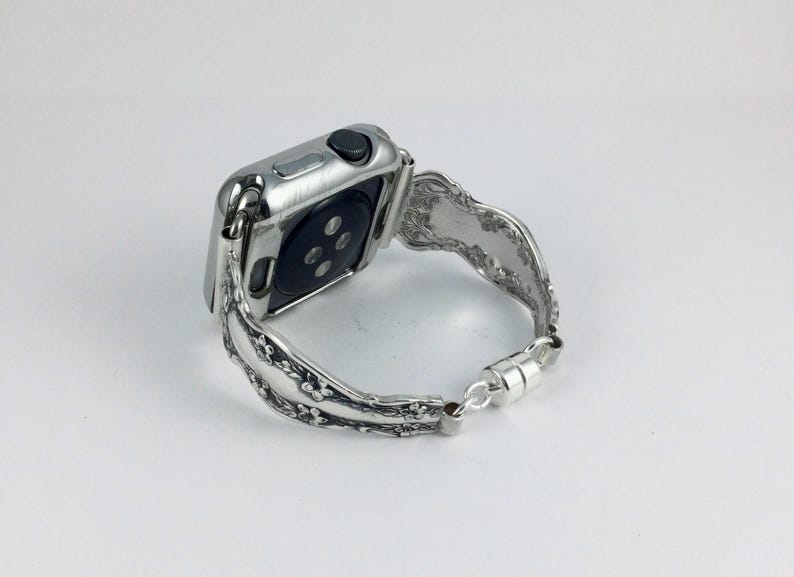 May include: A silver-toned smartwatch with a metallic, floral-patterned band. The watch features a rectangular case with a black circular sensor on the back and a side button. The band is connected to the watch case and secured with a magnetic clasp.