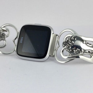 Puede incluir: Un reloj inteligente Fitbit plateado con una pantalla negra est&aacute; conectado a una pulsera plateada hecha de dos cucharas. Las cucharas est&aacute;n dobladas en forma de coraz&oacute;n y tienen dise&ntilde;os ornamentados.