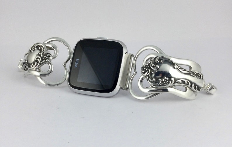Puede incluir: Un reloj inteligente Fitbit plateado con una pantalla negra est&aacute; conectado a una pulsera plateada hecha de dos cucharas. Las cucharas est&aacute;n dobladas en forma de coraz&oacute;n y tienen dise&ntilde;os ornamentados.
