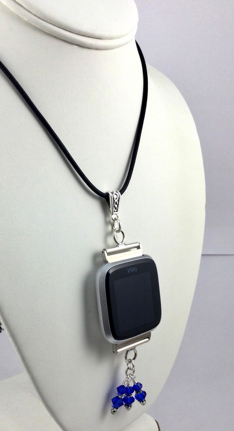 May include: A silver Fitbit smartwatch pendant necklace with a black cord. The watch has a black screen and is accented with a cluster of blue beads. The necklace is displayed on a white mannequin.