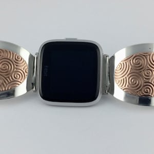 May include: A silver and rose gold watch band with a swirling pattern. The band is designed to fit a Fitbit watch.