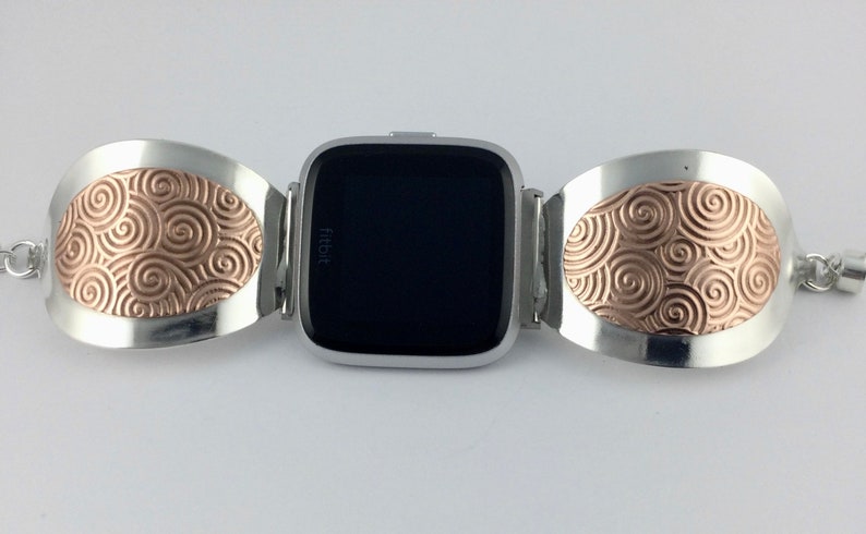 May include: A silver and rose gold watch band with a swirling pattern. The band is designed to fit a Fitbit watch.