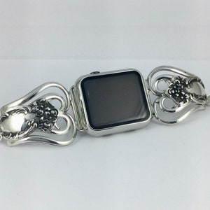 May include: Silver-toned Apple Watch band crafted from repurposed silverware. The band features ornate floral and scroll designs, with a central watch case and decorative spoon-shaped elements. A unique, handcrafted accessory.