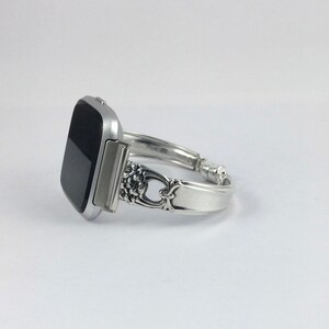 May include: A silver watch band made from a repurposed silverware piece. The band has a decorative floral design and is attached to a black square smartwatch.