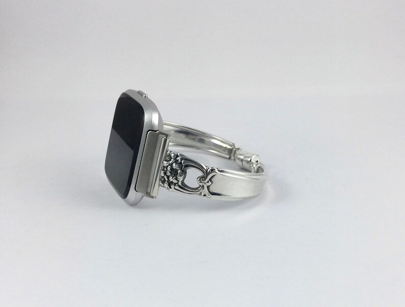 May include: A silver watch band made from a repurposed silverware piece. The band has a decorative floral design and is attached to a black square smartwatch.