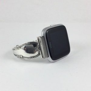 May include: A silver Fitbit smartwatch attached to a silver spoon bracelet. The watch face is black and displays the word "fitbit".